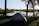 Photo of a green military tent pitched next to a calm lake with forest in the background under a clear sky.