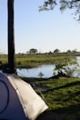 A tranquil campsite by a river with a tent set up under a tree. The background features a serene view of the river surrounded by lush green grass and scattered trees. The sunlight casts gentle shadows, creating a peaceful atmosphere.