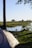 A tranquil campsite by a river with a tent set up under a tree. The background features a serene view of the river surrounded by lush green grass and scattered trees. The sunlight casts gentle shadows, creating a peaceful atmosphere.