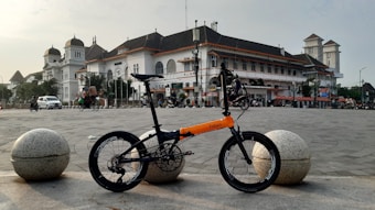 A folding bicycle with an orange and black frame is parked in an open square, with a large, historic building featuring a white facade and a dark roof in the background. The scene includes a few vehicles and people, with the sky partially cloudy.
