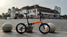 A folding bicycle with an orange and black frame is parked in an open square, with a large, historic building featuring a white facade and a dark roof in the background. The scene includes a few vehicles and people, with the sky partially cloudy.