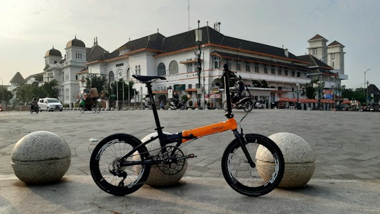 A folding bicycle with an orange and black frame is parked in an open square, with a large, historic building featuring a white facade and a dark roof in the background. The scene includes a few vehicles and people, with the sky partially cloudy.