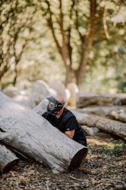 A person wearing a mask and protective gear is crouched behind a large fallen tree in a wooded area. The ground is covered with dry leaves and other logs are scattered around. Sunlight filters through the dense foliage, creating a dappled light effect.