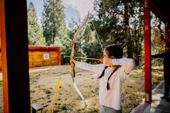 A young person with long hair and glasses is practicing archery outdoors. They are drawing a bow with a bright yellow arrow aimed at a distant target positioned on a hay bale. The setting is surrounded by tall trees under a clear sky.