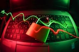 A laptop keyboard is illuminated by red and green light trails, with a padlock placed on top of the keyboard. The lighting creates a dynamic and somewhat futuristic atmosphere, emphasizing themes of security and technology.