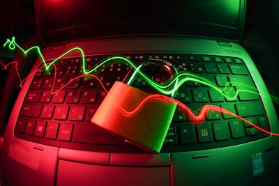 A laptop keyboard is illuminated by red and green light trails, with a padlock placed on top of the keyboard. The lighting creates a dynamic and somewhat futuristic atmosphere, emphasizing themes of security and technology.