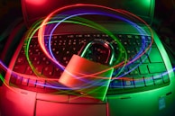 A padlock sits on a laptop keyboard with glowing red, green, and blue light trails swirling around, creating a sense of security and cyber awareness.