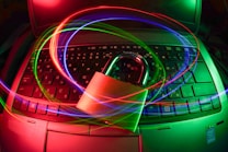 A padlock sits on a laptop keyboard with glowing red, green, and blue light trails swirling around, creating a sense of security and cyber awareness.