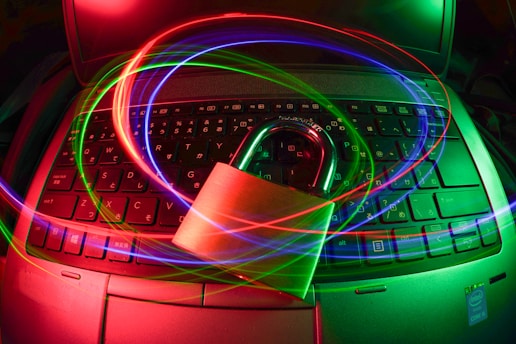 A padlock sits on a laptop keyboard with glowing red, green, and blue light trails swirling around, creating a sense of security and cyber awareness.