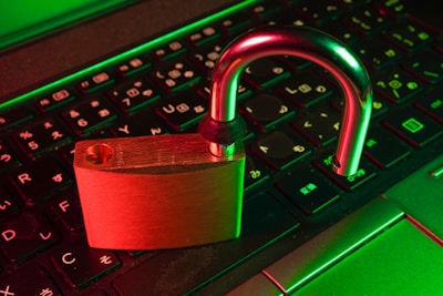 A brass-colored padlock is open, resting on the keyboard of a laptop. The keyboard has Japanese characters, and the scene is illuminated with green and red lighting, creating a dramatic contrast.