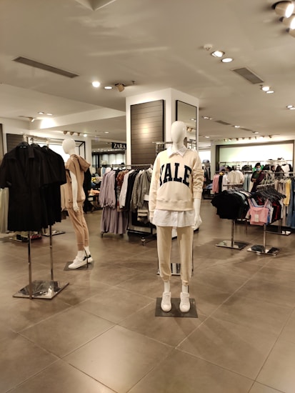 A clothing store interior with several mannequins displaying casual wear. The foreground mannequin is dressed in a cream-colored sweatshirt with 'YALE' printed on it, paired with matching pants and white sneakers. Garment racks filled with various clothing items are visible in the background along with store signage.