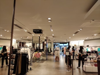 A retail clothing store interior features well-organized racks and shelves displaying various clothing items. Several people are browsing the selection. The lighting is bright and evenly distributed across the space, creating an inviting atmosphere. Mannequins are dressed in different outfits, showcasing the latest trends.