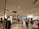 A retail clothing store interior features well-organized racks and shelves displaying various clothing items. Several people are browsing the selection. The lighting is bright and evenly distributed across the space, creating an inviting atmosphere. Mannequins are dressed in different outfits, showcasing the latest trends.