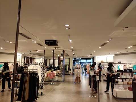 A retail clothing store interior features well-organized racks and shelves displaying various clothing items. Several people are browsing the selection. The lighting is bright and evenly distributed across the space, creating an inviting atmosphere. Mannequins are dressed in different outfits, showcasing the latest trends.