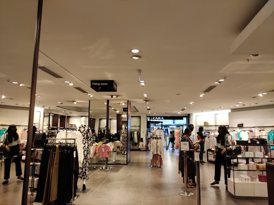 A retail clothing store interior features well-organized racks and shelves displaying various clothing items. Several people are browsing the selection. The lighting is bright and evenly distributed across the space, creating an inviting atmosphere. Mannequins are dressed in different outfits, showcasing the latest trends.