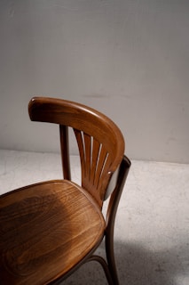 A wooden chair with a curved backrest and carved seat detail is positioned against a plain, light-colored wall. The chair's finish is a rich, warm brown, and the lighting casts soft shadows, creating a subtle play of light on the textured floor.
