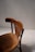 A wooden chair with a curved backrest and carved seat detail is positioned against a plain, light-colored wall. The chair's finish is a rich, warm brown, and the lighting casts soft shadows, creating a subtle play of light on the textured floor.