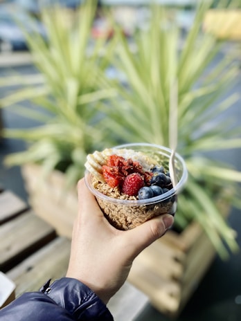 A hand holding a transparent plastic cup filled with an assortment of food, including granola, blueberries, strawberries, banana slices, and dried fruits. The background features blurred greenery and an outdoor setting, with a wooden surface partially visible.