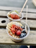 Two bowls of granola are on a wooden table. Each bowl is topped with sliced bananas, fresh blueberries, dried fruits, and chia seeds. Wooden spoons are placed in each bowl.