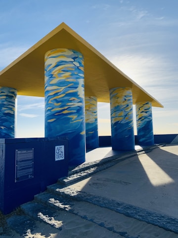 A structure with four large pillars painted in a swirling blue, yellow, and white pattern supports a yellow, flat roof. The structure casts long shadows on the ground. A nearby plaque on a blue wall has writing and the words 'Art Mongers' visible.
