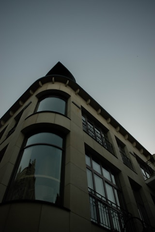 The image features an architectural perspective of a modern building with a curved facade. Large windows extend vertically, reflecting a hint of an adjacent structure. The design is sleek and contemporary, with visible geometric lines and a muted color palette.