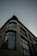 The image features an architectural perspective of a modern building with a curved facade. Large windows extend vertically, reflecting a hint of an adjacent structure. The design is sleek and contemporary, with visible geometric lines and a muted color palette.