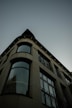 The image features an architectural perspective of a modern building with a curved facade. Large windows extend vertically, reflecting a hint of an adjacent structure. The design is sleek and contemporary, with visible geometric lines and a muted color palette.