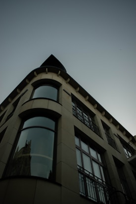 The image features an architectural perspective of a modern building with a curved facade. Large windows extend vertically, reflecting a hint of an adjacent structure. The design is sleek and contemporary, with visible geometric lines and a muted color palette.