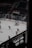 Hockey players dressed in red uniforms are on the ice, with one player in goal and others moving towards him. The ice rink has advertising boards, and there are empty seats in the darkened arena. A banner with dates is visible above the seating area.
