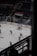 Hockey players dressed in red uniforms are on the ice, with one player in goal and others moving towards him. The ice rink has advertising boards, and there are empty seats in the darkened arena. A banner with dates is visible above the seating area.