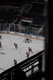 Hockey players dressed in red uniforms are on the ice, with one player in goal and others moving towards him. The ice rink has advertising boards, and there are empty seats in the darkened arena. A banner with dates is visible above the seating area.