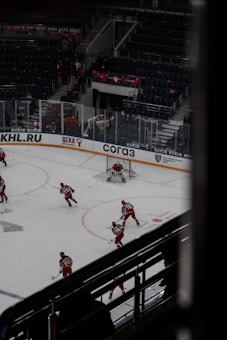 Hockey players dressed in red uniforms are on the ice, with one player in goal and others moving towards him. The ice rink has advertising boards, and there are empty seats in the darkened arena. A banner with dates is visible above the seating area.