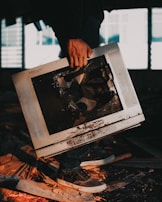 A person is holding a broken and dirty television set with a cracked screen. The surroundings appear to be a dimly lit, possibly abandoned space with debris and wooden planks on the floor. The lighting casts shadows, giving a dramatic effect.
