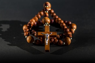 A wooden rosary with a cross at its center is positioned against a dark background. The beads are spherical and connected by a string, creating a circle around the cross. The figure of Jesus is depicted on the cross, adding a silver accent to the piece. Soft lighting casts shadows, enhancing the texture of the wood.