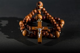 A wooden rosary with a cross at its center is positioned against a dark background. The beads are spherical and connected by a string, creating a circle around the cross. The figure of Jesus is depicted on the cross, adding a silver accent to the piece. Soft lighting casts shadows, enhancing the texture of the wood.