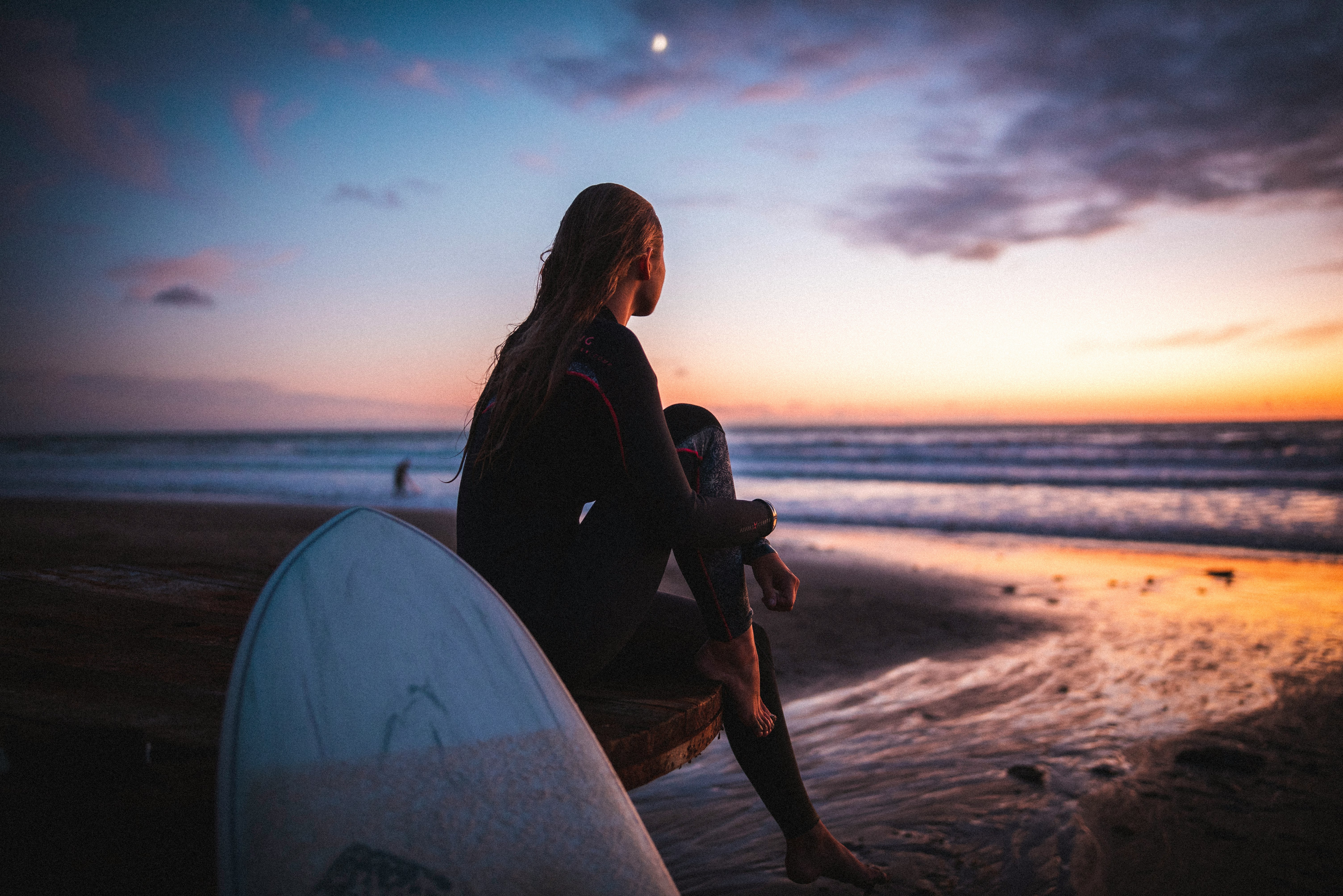 woman in black jacket sitting on white surfboard on beach during sunset