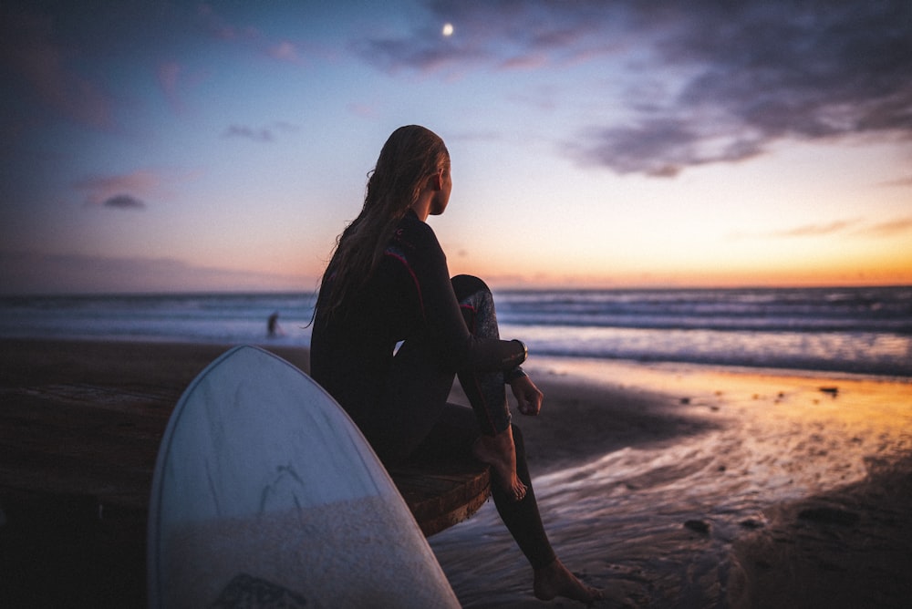 Mujer en chaqueta negra sentada en tabla de surf blanca en la playa durante la puesta del sol