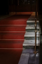 A set of stairs is covered with a red carpet leading up to a closed door. The stairs are flanked by a brass railing, and the steps are made of a mix of dark gray and white marble.