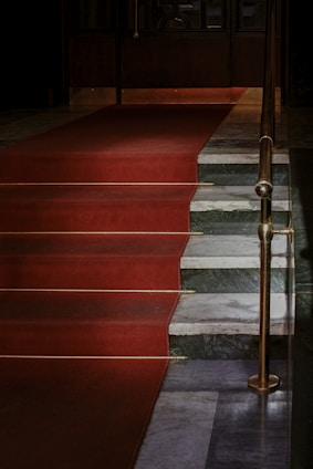 A set of stairs is covered with a red carpet leading up to a closed door. The stairs are flanked by a brass railing, and the steps are made of a mix of dark gray and white marble.