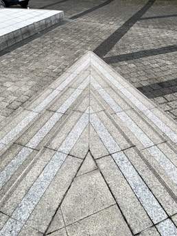 A geometric pattern formed by textured pavement with triangular and linear designs. The surface is made of arranged stone tiles, creating a visually intriguing perspective. Tension between textures and angles adds depth and motion.