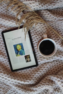 A cozy setup featuring a digital tablet displaying an article with an image, placed on a textured, woven blanket. Beside the tablet is a mug filled with a dark beverage, likely coffee. A sprig of dried grass lies across the scene, adding a natural element.