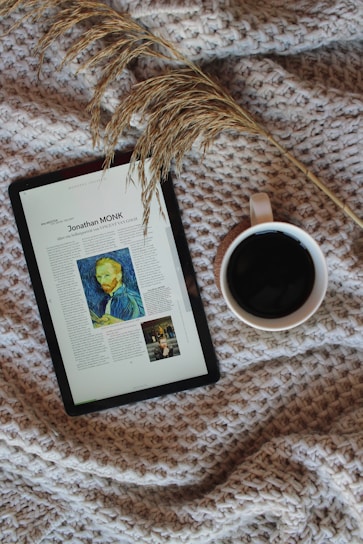 A cozy setup featuring a digital tablet displaying an article with an image, placed on a textured, woven blanket. Beside the tablet is a mug filled with a dark beverage, likely coffee. A sprig of dried grass lies across the scene, adding a natural element.