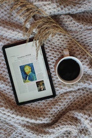 A cozy setup featuring a digital tablet displaying an article with an image, placed on a textured, woven blanket. Beside the tablet is a mug filled with a dark beverage, likely coffee. A sprig of dried grass lies across the scene, adding a natural element.