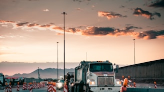 A large truck is driving through a construction zone at sunset, with mountains in the background. Traffic cones line the road, and construction workers are visible nearby. Streetlights are getting illuminated as the sky transitions from day to night.