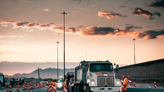 A large truck is driving through a construction zone at sunset, with mountains in the background. Traffic cones line the road, and construction workers are visible nearby. Streetlights are getting illuminated as the sky transitions from day to night.