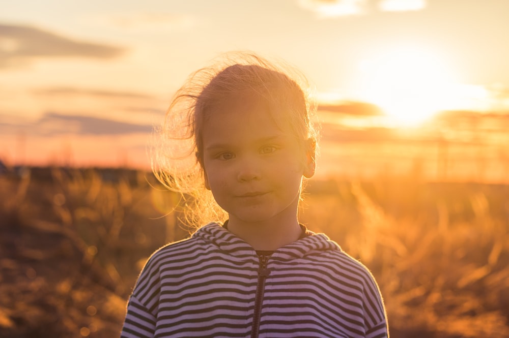 Muchacha en camisa de rayas blancas y azules de pie en el campo durante la puesta del sol