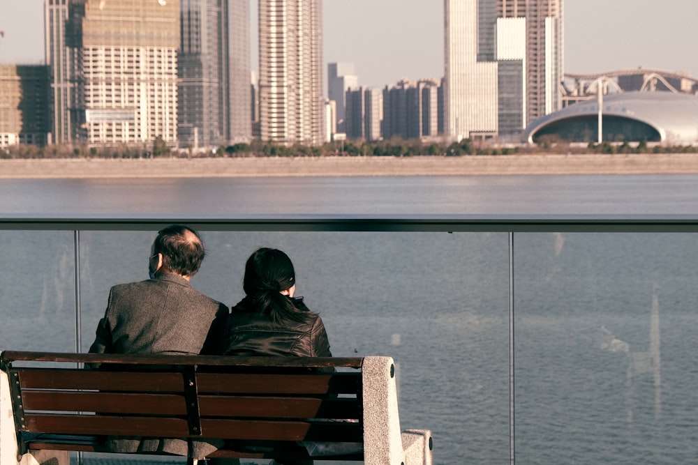 couple sitting on brown wooden bench near body of water during daytime