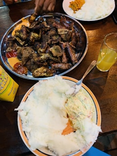 A table set with a large metal platter filled with grilled meat pieces surrounded by different sauces. Nearby is another plate with a large serving of a white starchy food similar to mashed potatoes or nsima, accompanied by a creamy salad. A glass filled with orange juice and a bottle of sauce are also visible.