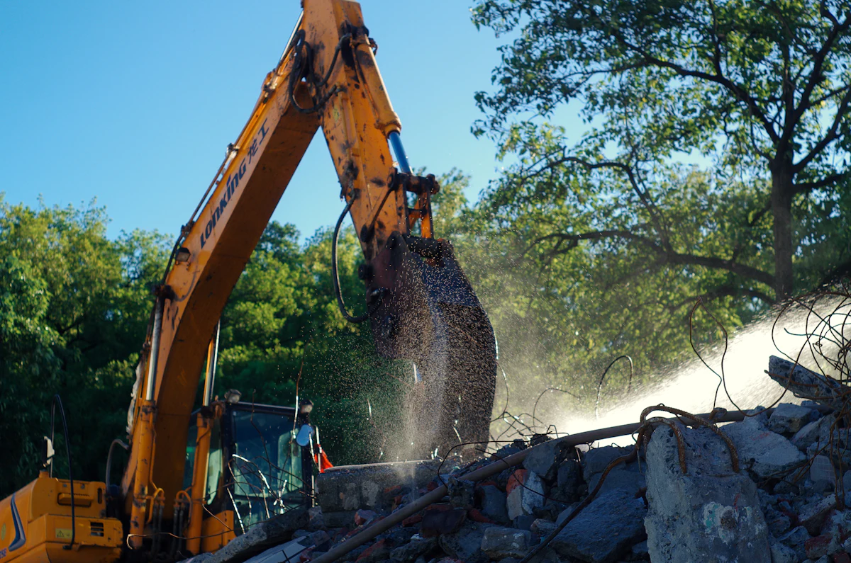 An excavator with a grapple attachment lifts debris from a rubble pile, dust drifting through the frame with trees behind.