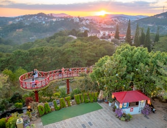 A scenic viewpoint overlooking a lush green valley with a vibrant sunset in the background. A circular viewing platform with a red railing is situated among the trees, where a few people are enjoying the view. Below, there is a small structure with a red roof, adorned with signs and lanterns. The foreground features a neatly arranged garden with letters spelling out 'DA LAT VIEW'. Houses are scattered on the distant hills, surrounded by dense forest.
